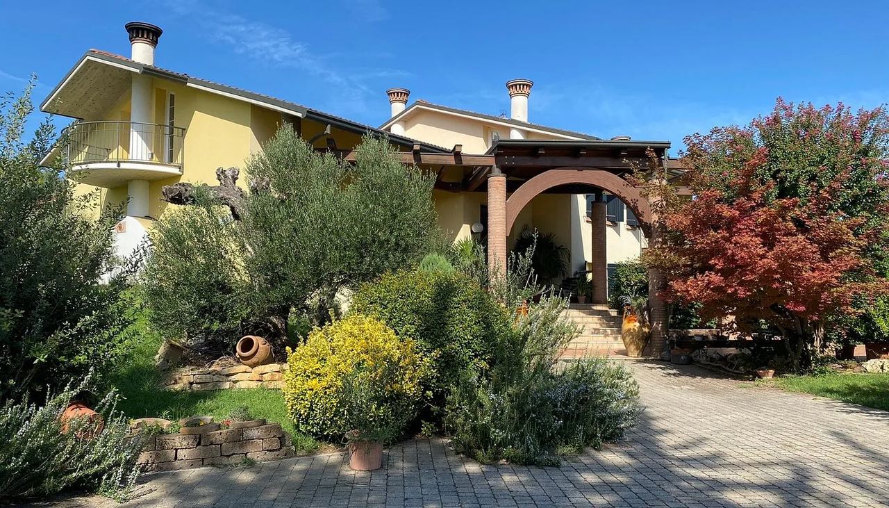 La villa vista dal giardino, con il pergolato d'ingresso e gli ulivi sotto il cielo azzurro